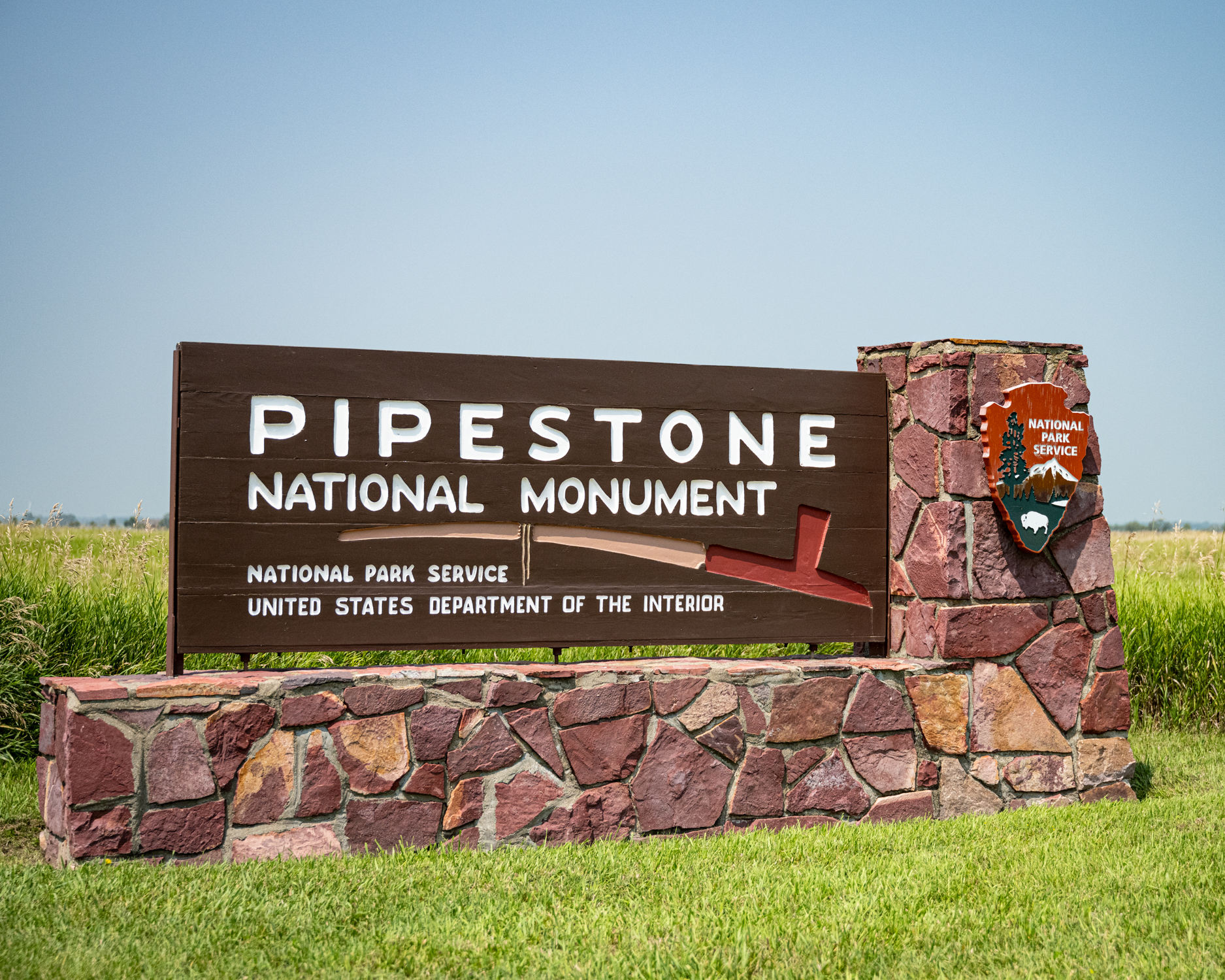 Pipestone Entrance Sign Image shows the entrance sign to Pipestone National Monument. It is a large brown sign with text reading: Pipestone National Monument, National Park Service, United States Department of the Interior. Across the center of the sign is a stylized image of a