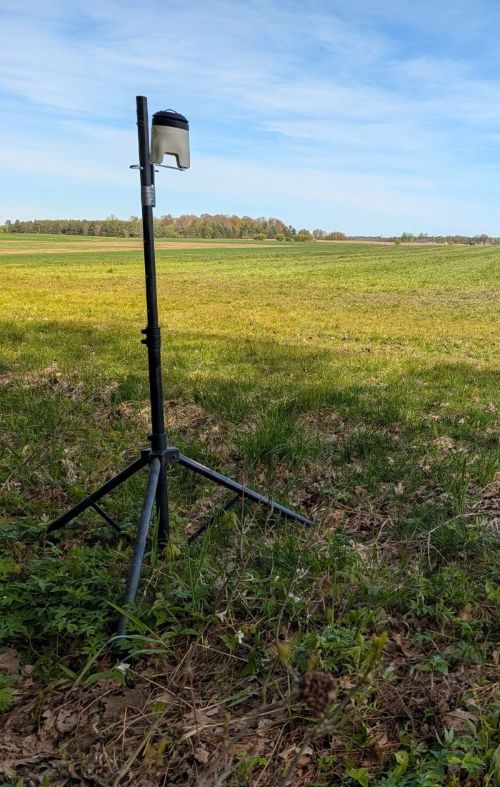 A black tripod sits on grass. At the top of the tripod is a football-sized device used to measure air quality.
