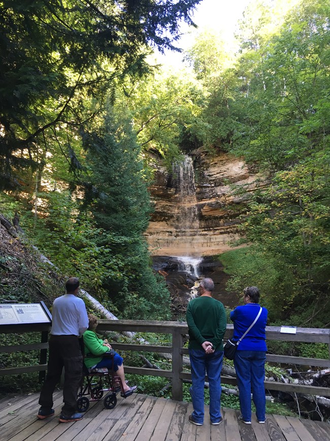 Four people admire a tall waterfall from a wooden platform
