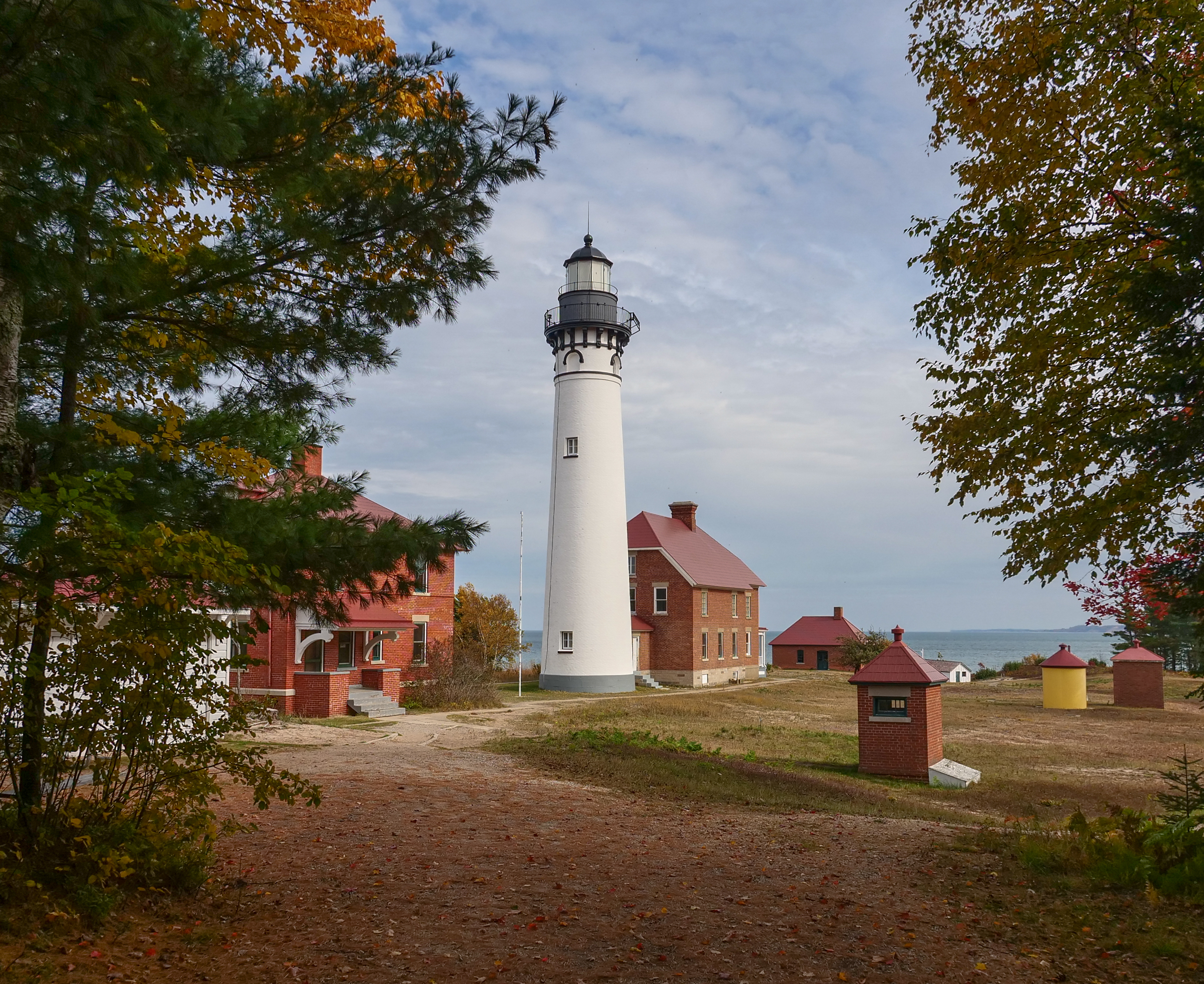 Au Sable Light Station 1874 A tall white lighthouse and red brick buildings are seen through a clearing in the trees.