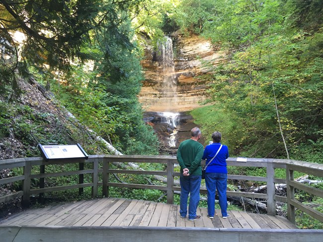 Two visitors admiring a tall waterfall in a wooded area.