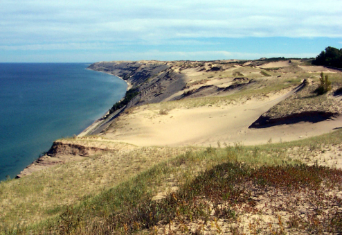 Wide sweeping sand dunes that rise high above the edge of a lake with some grasses growing on top.