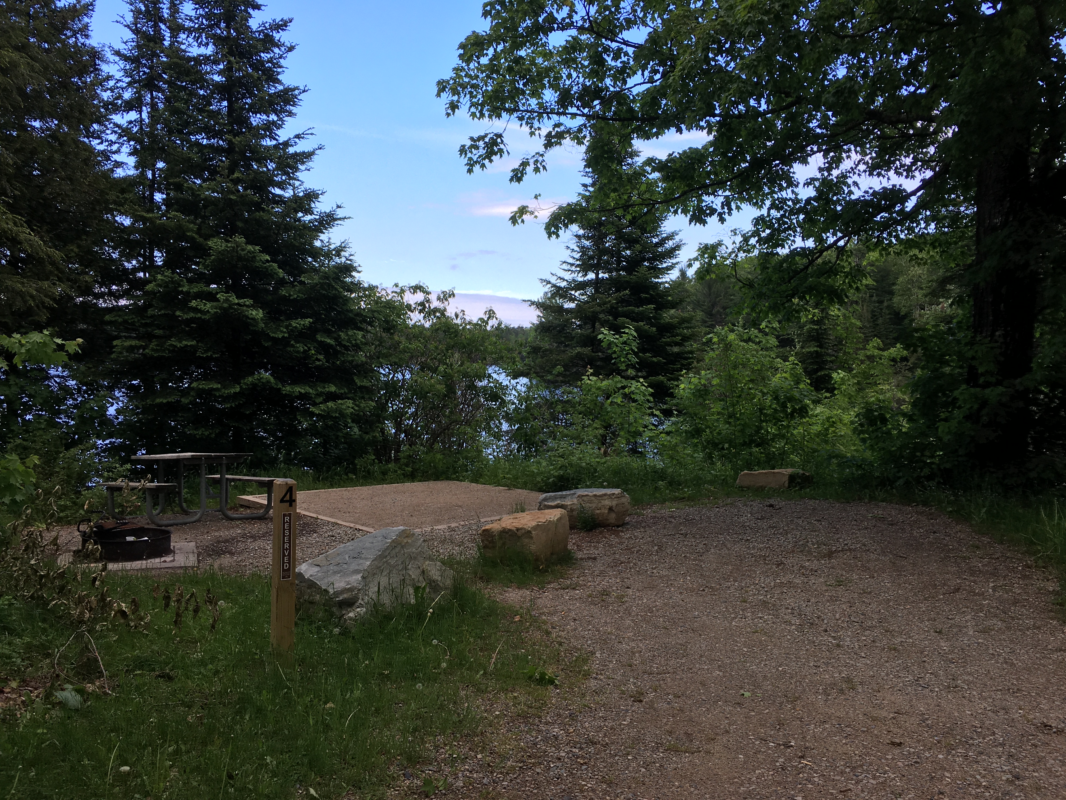 Gravel parking spot next to campsite pad, picnic table and grill on the edge of a lake.