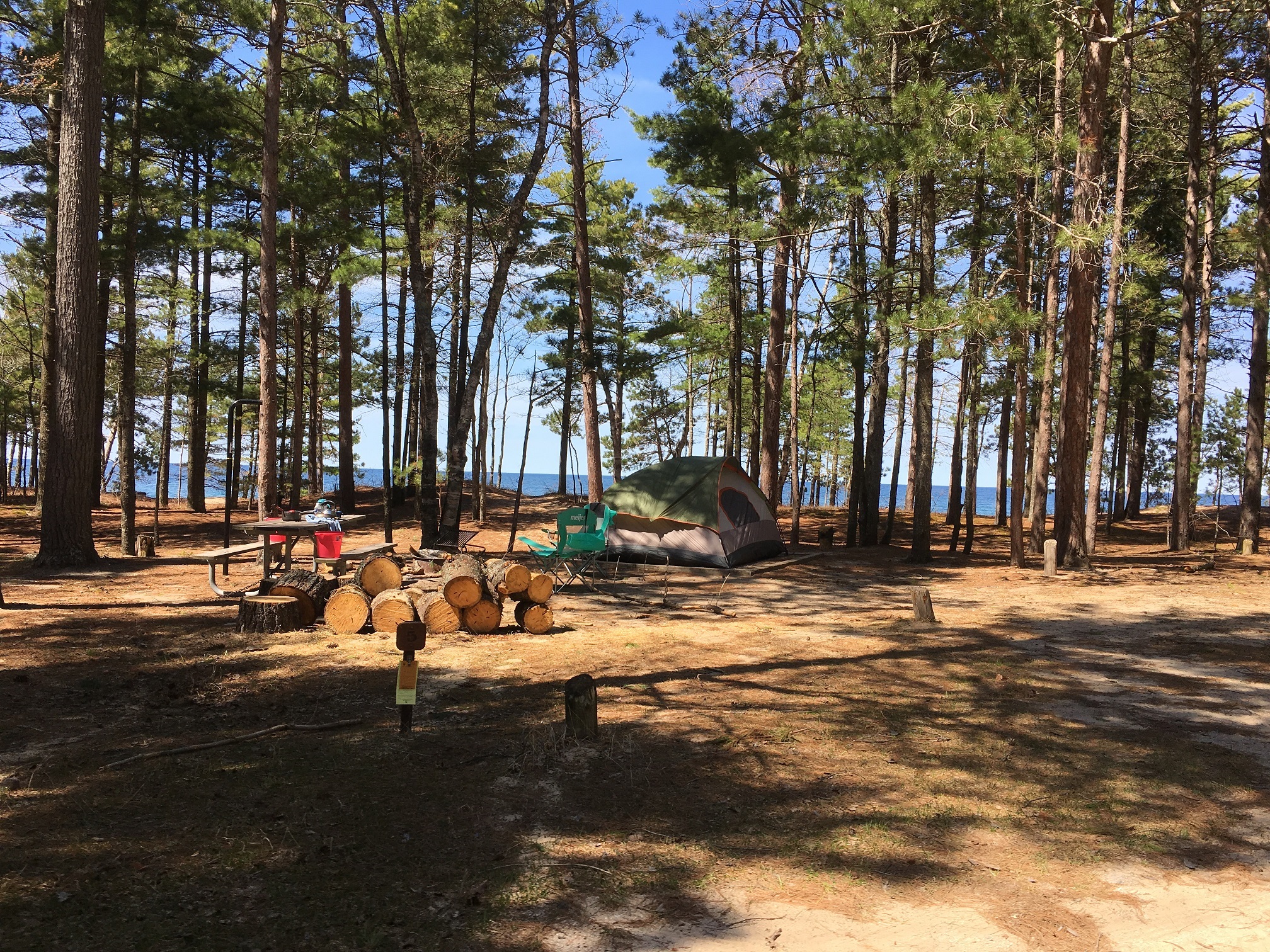 Campsite at Twelvemile Beach Campground surrounding by pine trees with Lake Superior in the background. Includes tent, chair, picnic table, and wood pile.