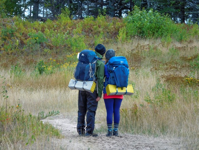 Two people with their backs to the camera looking at a phone. They have large packs on their back and sleeping pads attached to their packs.