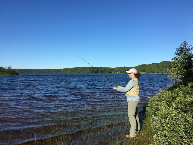 Woman standing on grassy water's edge while fishing at a lake.