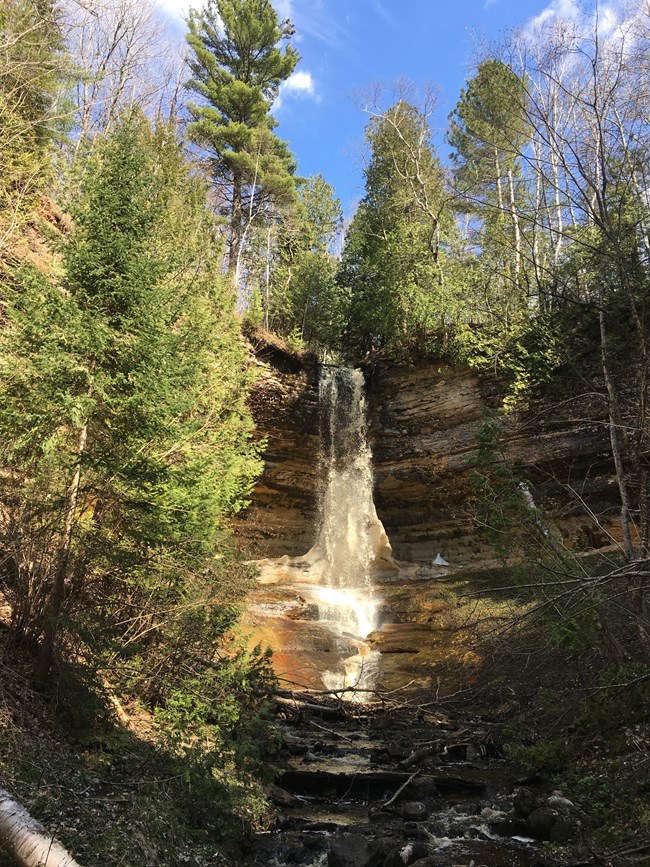 Waterfall flows from tree-topped sandstone cliff onto icy cone below.