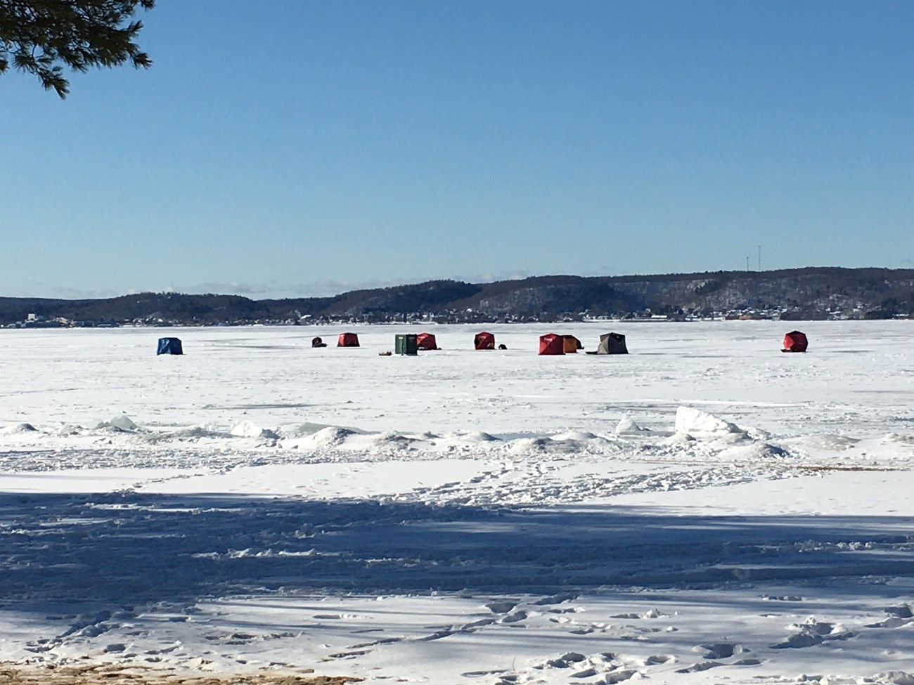 Eight fishing shacks on the ice of a large bay on a sunny day under a blue sky