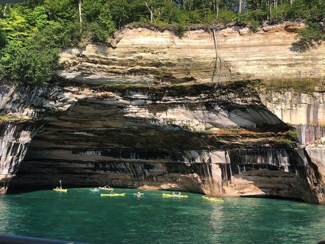 Group of yellow kayaks alongside a colorful concave section of the cliffs