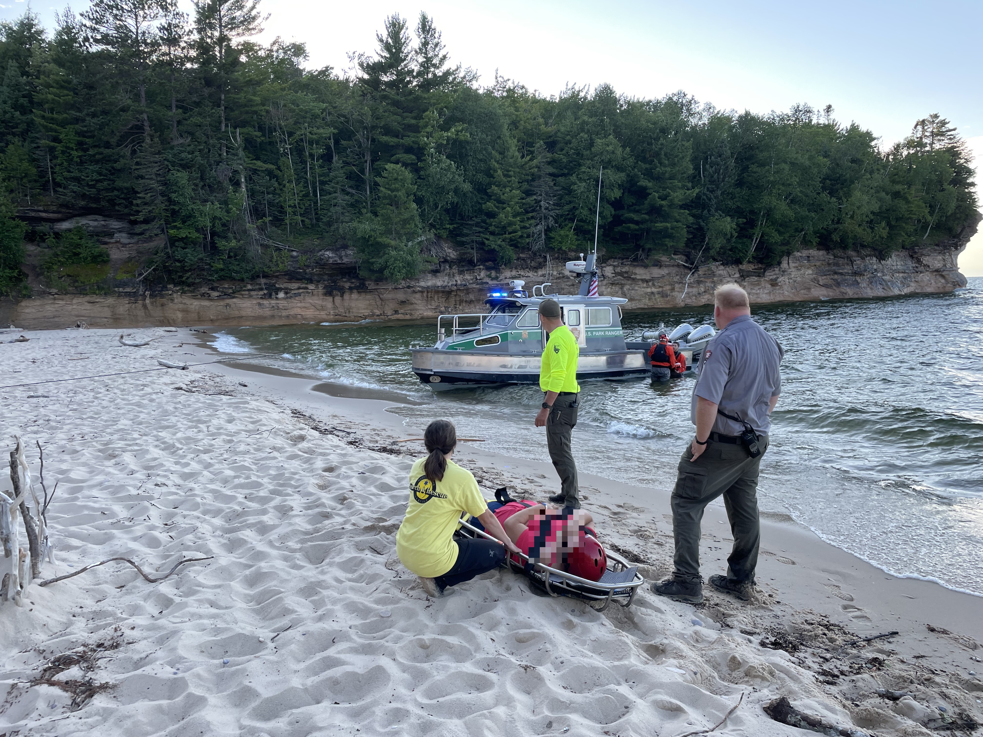 Several people surround a person on a litter on a beach. There is a boat pulled up to the beach.