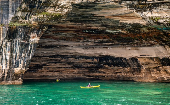 Two yellow sea kayaks next to towering mineral-stained sandstone cliffs.