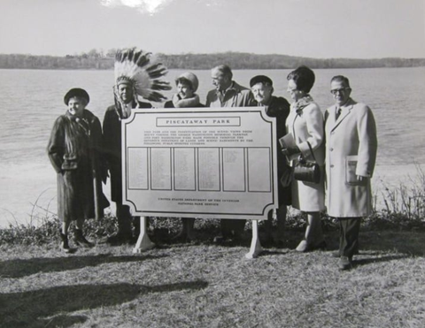Seven people stand behind a sign with the title 'Piscataway Park'. One man wears a large feather headdress. A river flows behind the group.