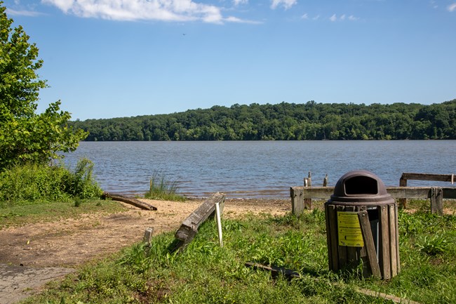 A wood railing and trash can at the edge of a riverbank with a dirt path leading to the water.