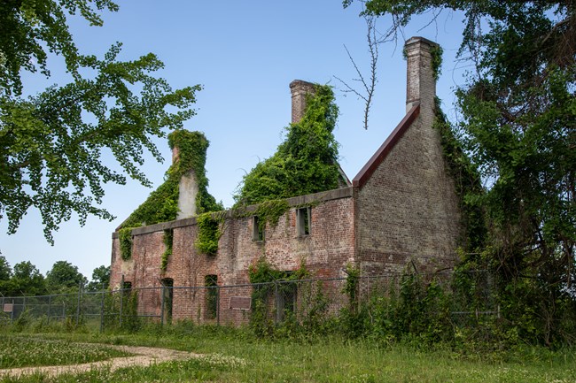 Ruins of a rectangular brick building without a roof covered in vines.