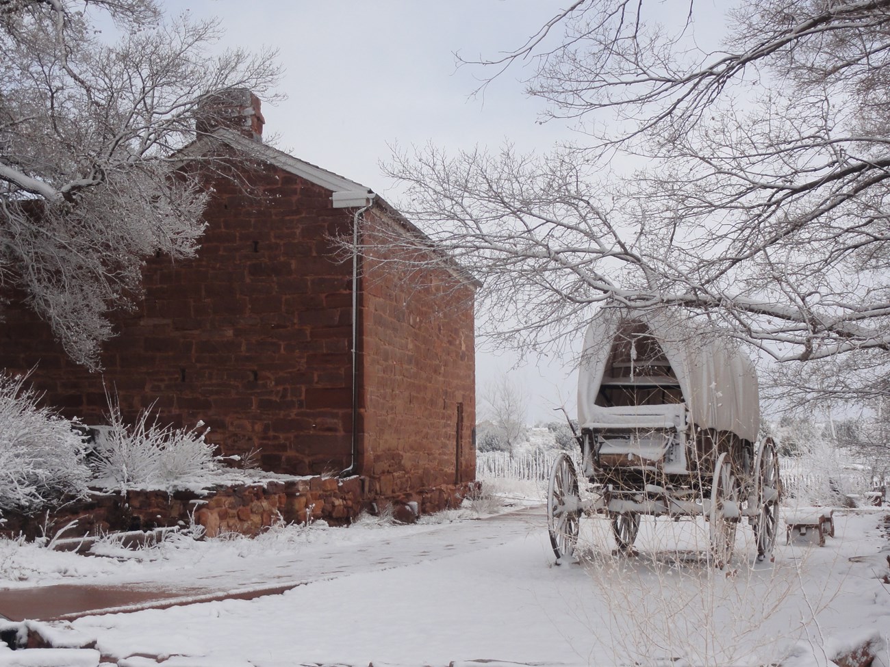 Snow has fallen on a two story red stone building next to a covered wagon.