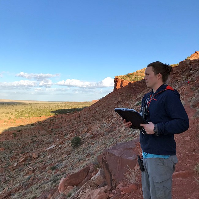 A man stands on a red-rock hillside, wearing binoculars and holding a clipboard.