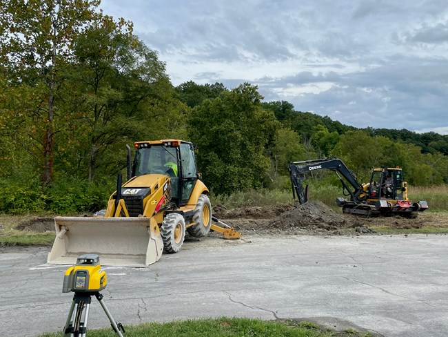 Parking lot with a yellow bulldozer and digger, digging up the dirt area behind the dozer. The background it filled with green leaved trees.