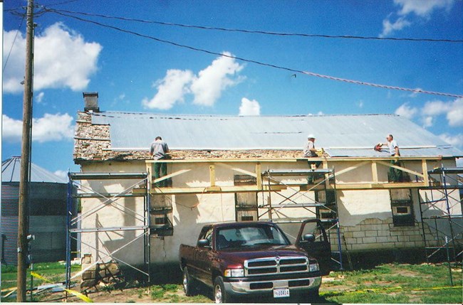 Three people stand on scaffolding in front of a white stucco building with 4 boarded windows and metal sheets over a wooden roof. A dark red truck with a door open is parked under the scaffolding. The sky is bright blue with a few clouds.