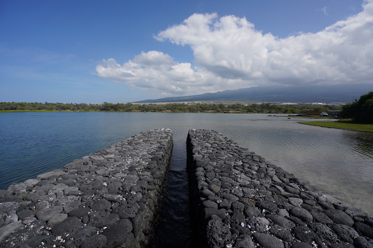 A water channel built with water worn lava rocks facing towards a fishpond. Blue skies with clouds and Hualalai volcano in background.
