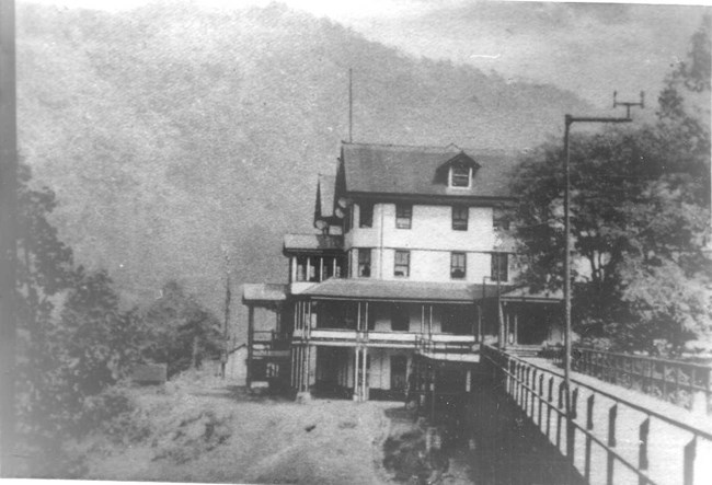 A black and white photo of a three sided wooden building with wrap around porch sits by the riverside