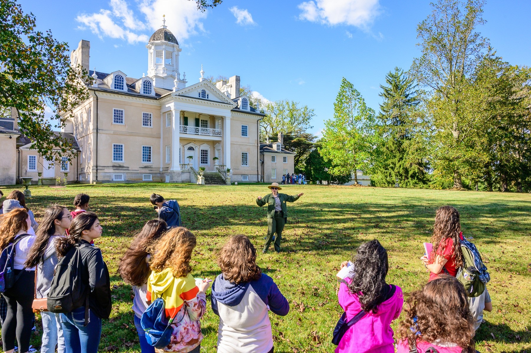Students with their backs turned, face a uniformed park ranger who is gesturing to a large pale yellow house in the background.