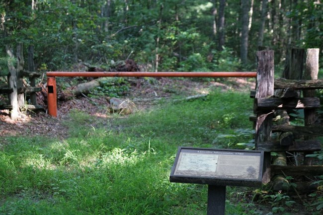 A wayside exhibit is to the right of a grassy trail with a closed gate.