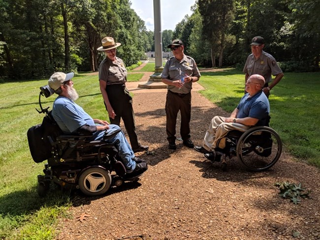 Two men in wheelchairs meet with Park Rangers on park trail