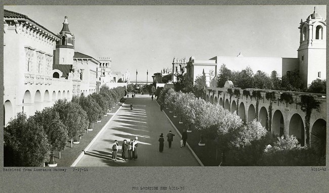 black and white photograph of people in dresses and suits walking along a stone path lined with trees and buildings with arches.