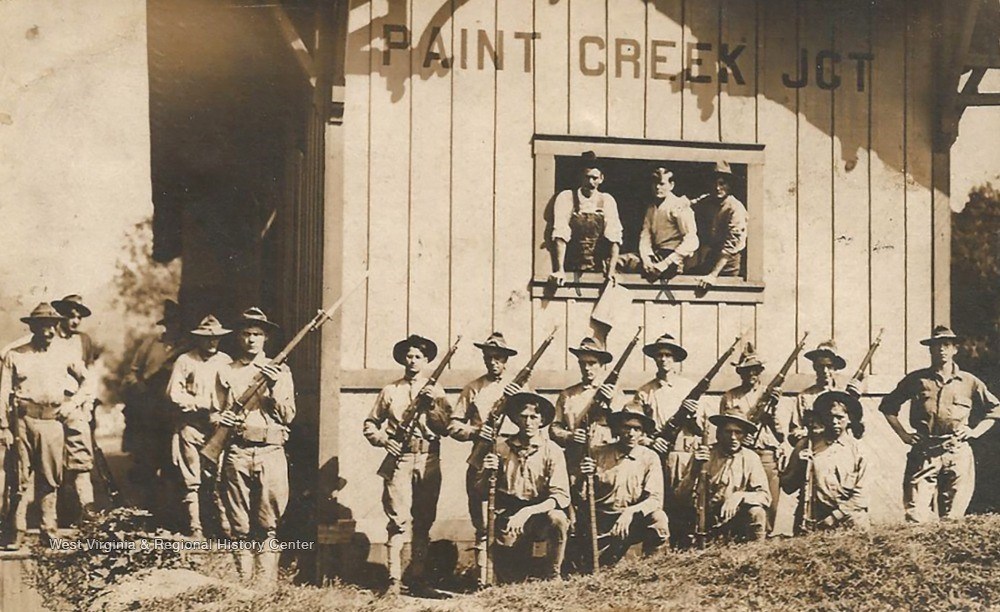 A row of men in white shirts and hats stand near a wooden shack holding rifles. On the shack wall above the men is a painted label reading Paint Creek Jct.