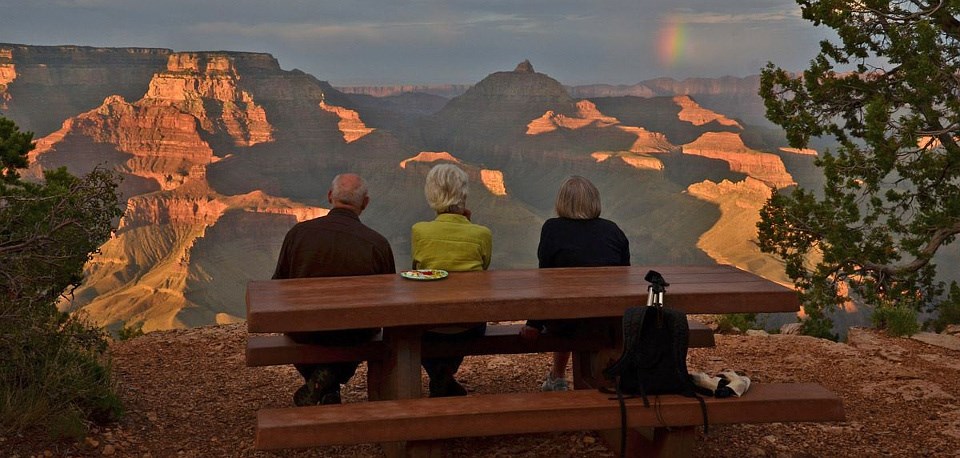 Three people sitting at a picnic table watching sunset and a rainbow over the Grand Canyon