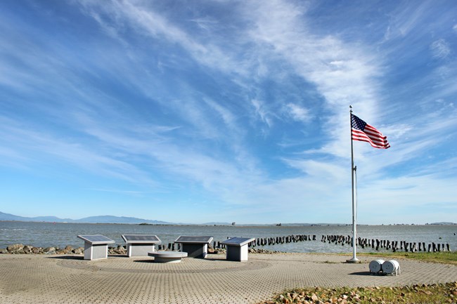 Four granite memorial stones are placed in an arc. An american flag is raised on the right. Bay water in the background.