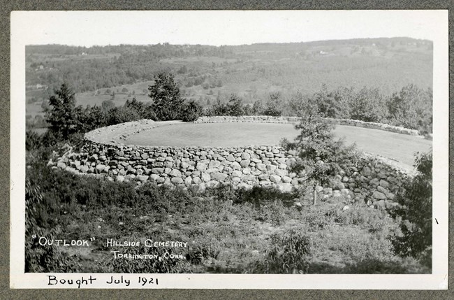 Black and white of flat grassy area circled with rocks on top of hill, with trees and shrubs planted around and views of the hills in the distance.