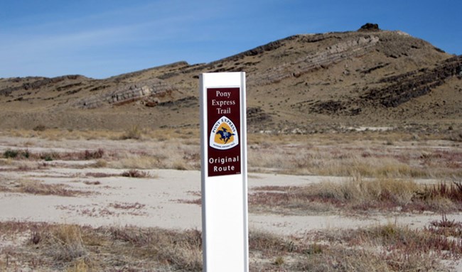A white post with a brown label on it stands in front of a brown rock hill with grass and dirt in the background. A white post with a brown label on it stands in front of a brown rock hill with grass and dirt in the background.