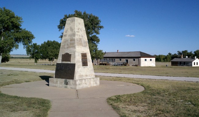 A large concrete pillar shaped monument with bronze plaques on it stands in a paved circle with a path leading to it and buildings in the background. A large concrete pillar shaped monument with bronze plaques on it stands in a paved circle with a path leading to it and buildings in the background.