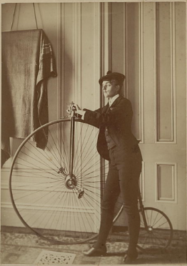 Woman poses humorously dressed as a man with a bike that has a large front wheel and small rear wheel