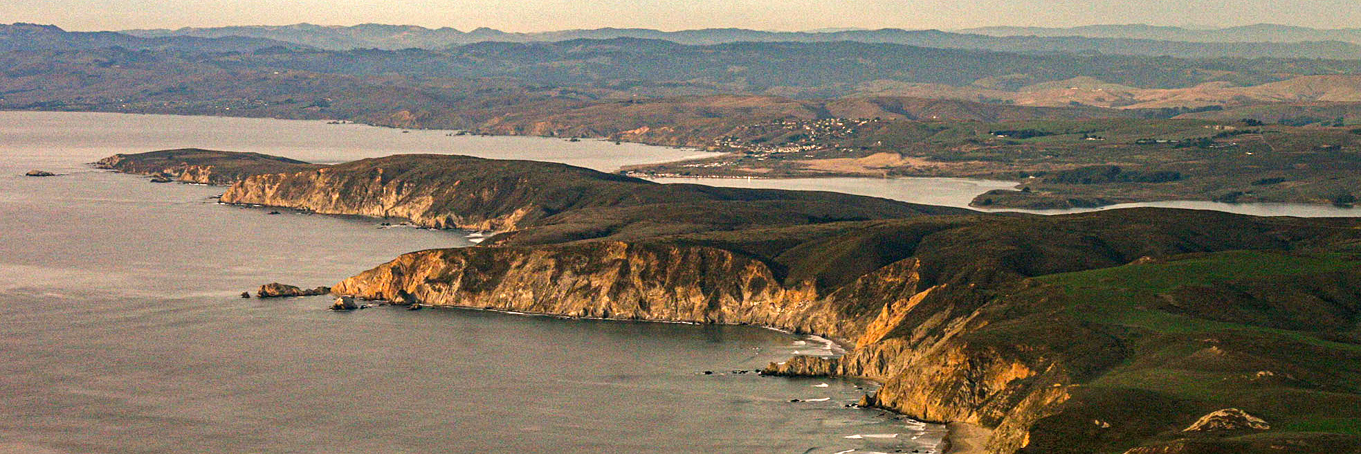 An aerial photo of a narrow grass-covered peninsula with beige cliffs rising above the ocean in the lower left. A narrow bay separates the peninsula from the mainland, which stretches into the distance.