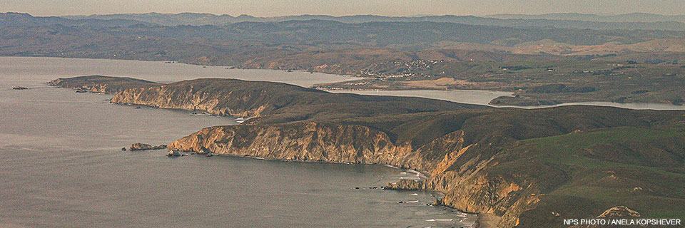 An aerial photo of Tomales Point taken on November 27, 2014. An aerial photo of a narrow grass-covered peninsula with beige cliffs rising above the ocean in the lower left. A narrow bay separates the peninsula from the mainland, which stretches into the distance.