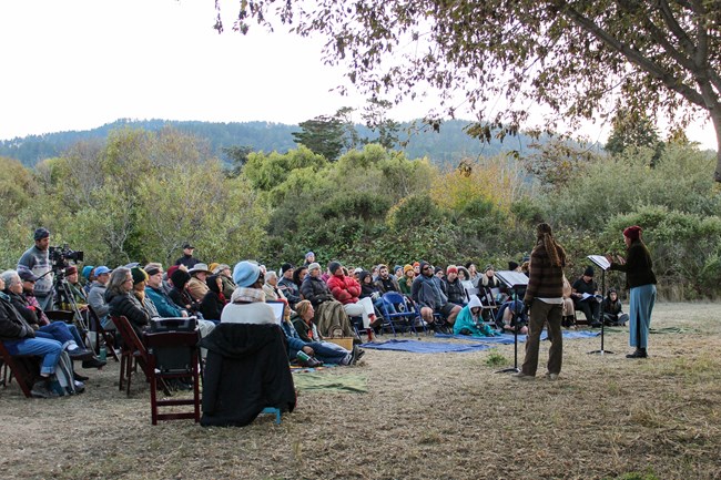 Two individuals read from material on musical stands in front of an audience of about 50 people sitting outdoors.