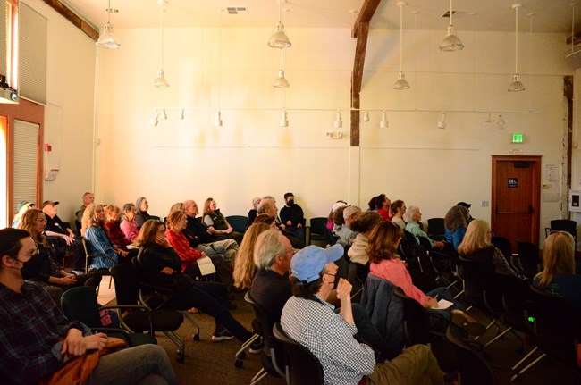 Thirty-six people sit facing to the right in a white-walled room.