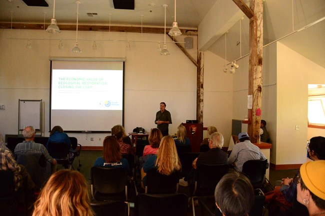 A man stands in front of a seated audience and adjacent to a screen on which a slide with a title of "The Economic Value of Ecological Restoration: Closing the Loop" is projected.