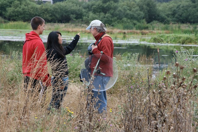 A man holding a large parabolic microphone stands next to two teenagers on the edge of a wetland.
