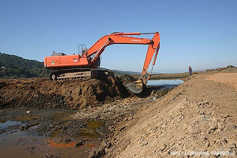 A large, orange excavator removes a section of a levee as water starts to flow through the breach.