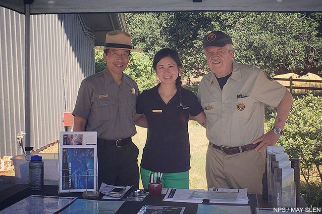 A ranger, a Community Volunteer Ambassador, and a volunteer stand together behind a table under a canopy.