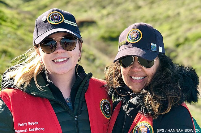 Winter Wildlife Docents Hannah Rouley and Michelle Van Aalst at the Elephant Seal Overlook on February 24, 2018. Two women wearing red volunteer vests and gray volunteer ball caps stand side-by-side with a grassy hillside rising behind them.