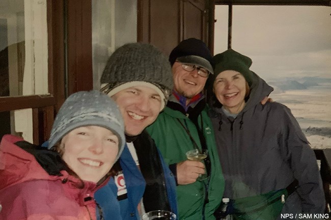 Two men and two women wearing warm clothing stand on an outside deck of a fire lookout with gray clouds and a valley floor in the distance.