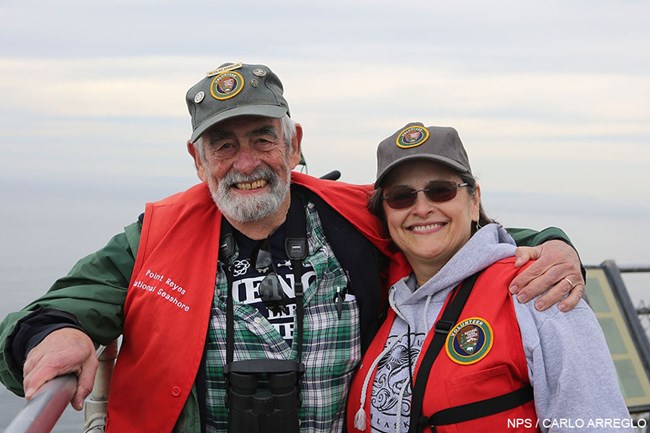 Winter Wildlife Docents Nik Epanchin and Tori Norman at the Lighthouse Observation Deck, on January 7, 2018. A gray-bearded man and a woman wearing red volunteer vests and gray volunteer ball caps stand close together with the ocean and gray skies in the background.