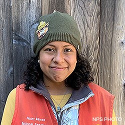 A head photo of Point Reyes Intern Ada wearing a red volunteer vest and a dark green Smokey Bear beenie hat.