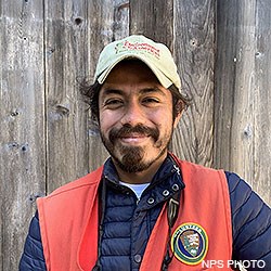 A head photo of Point Reyes Intern Kevin wearing a red volunteer vest over a blue jacket and a light green ball cap.