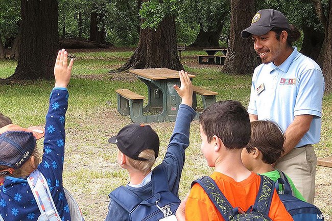 Children, some with hands raised, face a young Latino man in a picnic area.