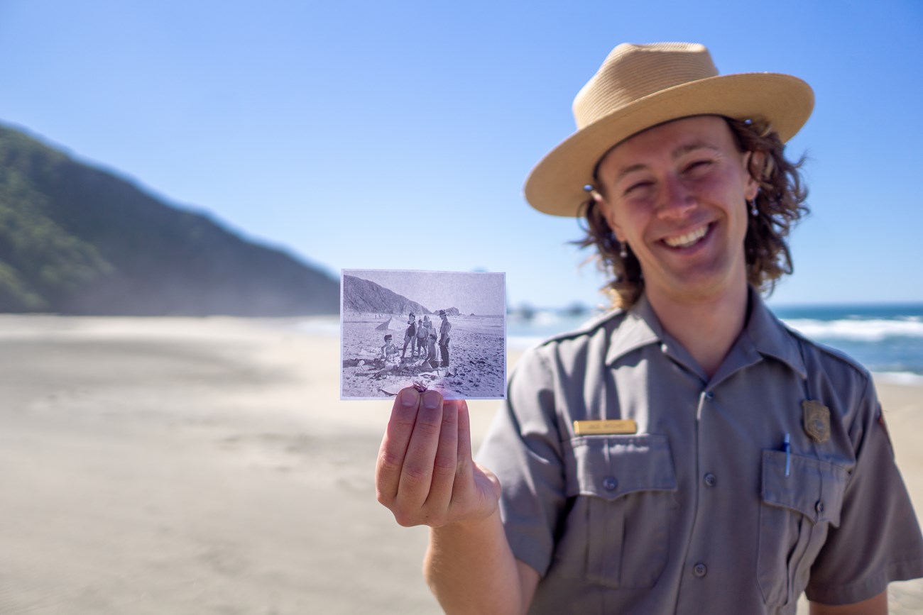 A smiling park ranger holds up a black and white photo on the beach.